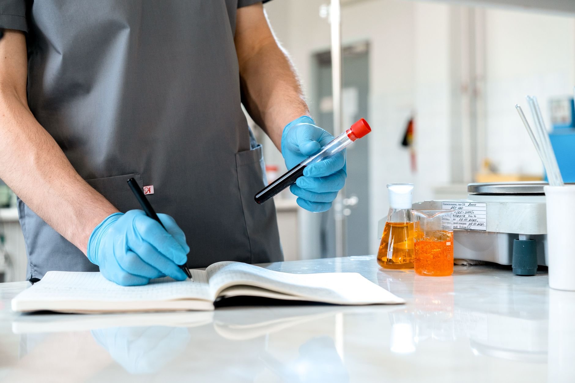 Scientist wearing gloves records lab observations, holding a test tube and writing in a notebook.